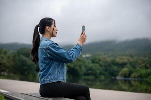 A woman is sitting on a bench by a stunning lake, using her smartphone to take a photo of the view.