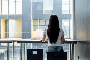 A rear view of an Asian woman sitting at a table by the window, working remotely from a coffee shop. photo