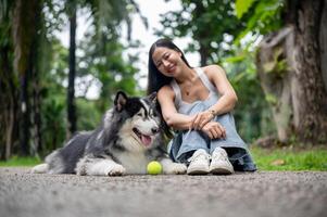A woman smiles at the camera while sitting on a street in a park, with her dog lying nearby. photo