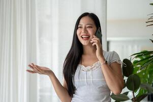 An attractive Asian woman is enjoying talking on the phone while standing by the window indoors. photo