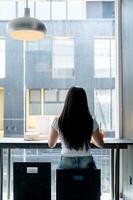 A rear view of an Asian woman sitting at a table by the window, working remotely from a coffee shop. photo