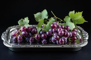 Fresh purple grapes garnished with green leaves on a silver tray against a dark background photo