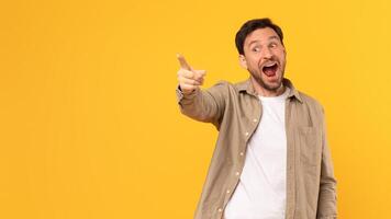 A man in a brown shirt stands in front of a yellow background, pointing his index finger at copy space and looking excitedly in that direction, with his mouth open in a smile. photo