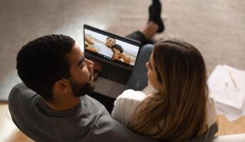 A couple is sitting on a couch at home, chatting with their friends on a laptop. The computer is open on a table in front of them, showing a call with two friends. photo