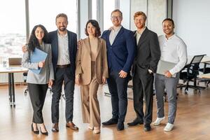 A group of six colleagues stand in a modern office, smiling, looking at the camera. They are a diverse group of professionals in business attire, demonstrating a company culture that values inclusion. photo