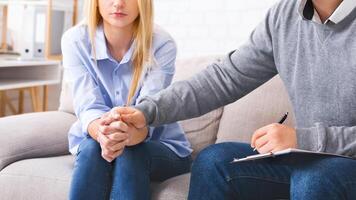 Cropped of woman is seated on a couch, clutching her hands nervously while engaging in a therapy session. Beside her, a man therapist is attentively listening and taking notes, touching client hand photo