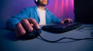 A close-up view of a gamers hand gripping a wired computer mouse, with the keyboard and a blurred figure in the background. The scene is lit by purple and pink lighting, cropped photo