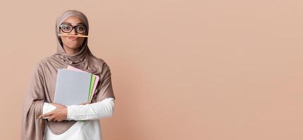 Black islamic female student in hijab keeping pencil like mustache and holding notebooks, fooling, having fun while posing over yellow background photo