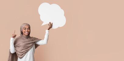 Idea Concept. Joyful black muslim woman in hijab holding empty speech bubble and pointing index finger up over yellow background, copy space photo