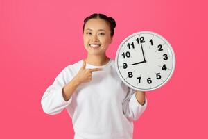 Asian woman with a bright smile holds a white clock in front of her on a pink background. She is wearing a white sweater and pointing at the clock with her right hand. photo