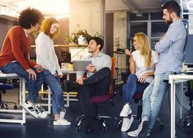 A group of four young professionals engage in a lively discussion in a brightly lit office space. One man sits on a chair holding a tablet, while the others stand around him, conversing and gesturing photo