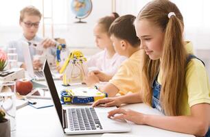 A young girl sits at a table in a classroom, using a laptop. She is surrounded by other students who are also working on projects. All kids focused on their work and appear to be enjoying themselves photo
