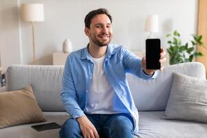 A man is seated on a couch, holding a smartphone in his hand. He appears focused on the screen, possibly browsing or texting. The room is cozy, with a soft light illuminating the scene. photo