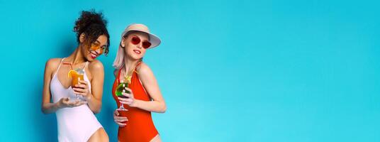 Two women wearing swimsuits and sunglasses are smiling and holding cocktails in front of a bright blue background. They appear to be enjoying themselves on a summer day, copy space photo