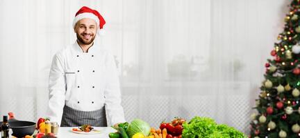 New Year Menu. Chef Man In Santa Hat Smiling At Camera Standing Next To Work Table In Kitchen. Selective Focus photo