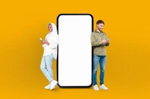 European man and black woman in hijab standing side by side, both looking at a cell phone in their hands, smartphone with white blank screen between them photo