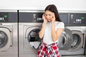 A young woman stands in a laundromat with her hands on her temples, looking stressed. She is dealing with unexpected laundry issues while several machines operate in the background. photo