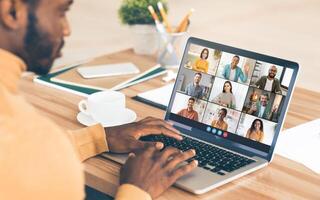 Black man sits at a desk in front of a laptop, participating in a virtual meeting with a group of people on screen. He is focused on the screen, his hands resting on the keyboard. photo