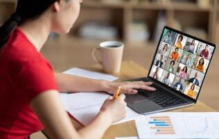 A woman sits at a desk with a laptop open in front of her. She is taking notes in a notebook with a pencil while attending an online meeting. The laptop screen displays a conference photo