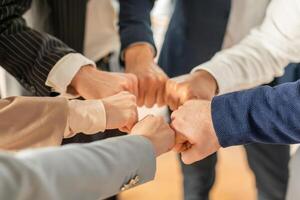 Several people, likely colleagues, stand in a circle and bump fists together. They are wearing business attire, suggesting they are in a professional setting, cropped photo