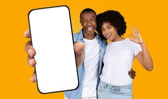 Black man and woman stand in front of yellow background. The man is holding a smartphone with a blank screen. The woman is making the ok sign with her hand. They both have big smiles on their faces. photo