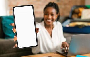 Black woman is seen holding up a cell phone in front of a laptop screen. She appears to be engaged in a call or browsing content on the phone while using the laptop simultaneously. photo