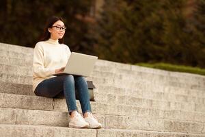 Korean woman is seated on steps outdoors, actively using a laptop computer. She appears focused on the screen, with her hands on the keyboard. The setting is urban, with buildings in the background. photo