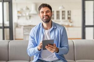 A man sits on a gray couch in his home and smiles while holding a tablet computer in his hands, websurfing or scrolling photo
