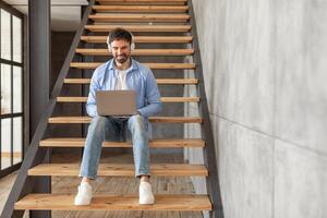 A man sits on wooden stairs in a modern home. He is wearing a blue shirt and jeans and is looking down at a laptop in his lap. He is smiling and has headphones on. He appears to be working photo