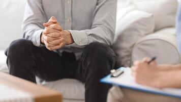 A man sits on a couch with his hands clasped, speaking with a therapist who takes notes on a clipboard during a session in a cozy living room setting, cropped photo