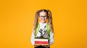 Smiling Schoolgirl Holding Book Stack And Microscope On Yellow Studio Background. Back To School Concept. Empty Space photo