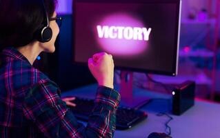 A gamer celebrates their victory in a dimly lit room with neon lighting. They have headphones on and are pumping their fist in the air. The word victory is displayed brightly on computer monitor. photo