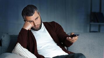 A man with a beard sits on a couch in a dimly lit living room, holding a TV remote with a bored expression on his face. He rests his head on his hand, leaning on a pillow photo