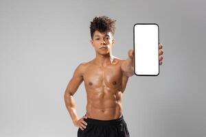 A young man stands confidently with a toned physique, showcasing a blank smartphone to the camera. His athletic stance emphasizes strength, while the neutral background photo