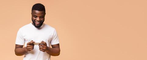 African American man with a beard, wearing a white t-shirt, smiling and using his smartphone. He is standing in front of a beige background. The man is looking down at his phone, copy space photo