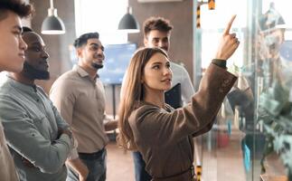 Portrait of young businesswoman mentor coach leader writing idea or task pointing at sticky notes on glass wall, diverse team developing work plan in creative corporate office at stand up meeting photo