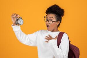 A young woman in a white sweatshirt and glasses stands in front of a bright yellow background with a surprised expression on her face as she holds up an alarm clock in one hand photo