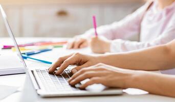 Woman is typing on a laptop computer while sitting at a desk. There are colored pencils and a notebook on the desk. Child is sitting in the background, drawing with a pink marker, cropped photo