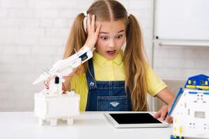 A young girl with pigtails looks surprised and concerned as she works on a robotics project. She is surrounded by robot parts and a tablet, suggesting she is following instructions photo