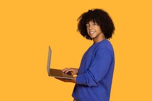 This image features a smiling young man with curly hair, wearing a blue sweatshirt, using a laptop computer against a bright yellow background photo