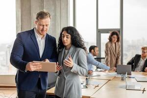 A man and a woman in business attire are discussing a project while looking at a tablet in a modern office setting. They are standing in the foreground with other colleagues working in the background photo