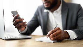 African American employer using smartphone and taking notes while working in office, selective focus on hand photo