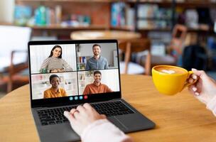 A person sits in a cafe, using a laptop to conference with three colleagues. They are smiling and engaged in conversation, enjoying a cup of coffee. The cafe is cozy and relaxed photo