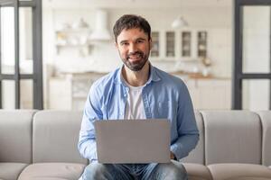 A man is sitting on a couch in a modern home, using a laptop computer. He is smiling and looking directly at the camera. The background is blurred and shows a light-colored living room photo