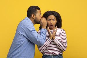 African American man dressed in a blue shirt is whispering into the ear of a young woman who looks surprised and shocked, isolated on yellow background photo