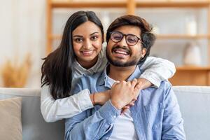 A joyful Indian couple embracing and smiling while sitting on a comfortable sofa in a warmly lit living room. They enjoy a moment of togetherness, creating a heartwarming, relaxed atmosphere. photo