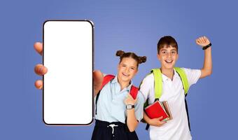 Two smiling students, a boy and a girl, pose together in front of a blue background, both have backpacks on and are holding books, showing phone with white blank screen photo
