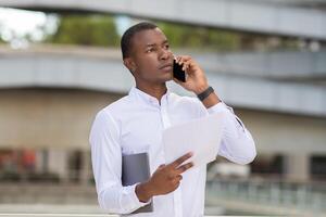 A young African American man stands in an urban environment, engaged in a phone conversation. He appears focused as he holds several documents in one hand and a tablet in the other. photo