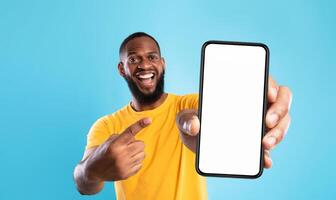 Mobile advertisement. Overjoyed black man pointing at cellphone with empty white screen on blue studio background, mockup for app or website. Cellphone display template photo