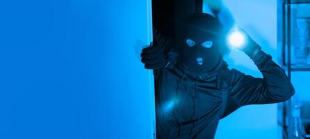 A person dressed in dark clothing and a ski mask peers into a dimly lit room while holding a flashlight, suggesting an illicit entry at night. The atmosphere is tense and foreboding. photo